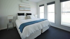 A hotel room with a large bed, two bedside tables, a wall-mounted air conditioner, and large windows with blinds.