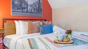 Une chambre d’hôtel avec un lit, des oreillers, une couette, un tableau encadré au mur et une petite table sur laquelle se trouvent une tasse et des fleurs.