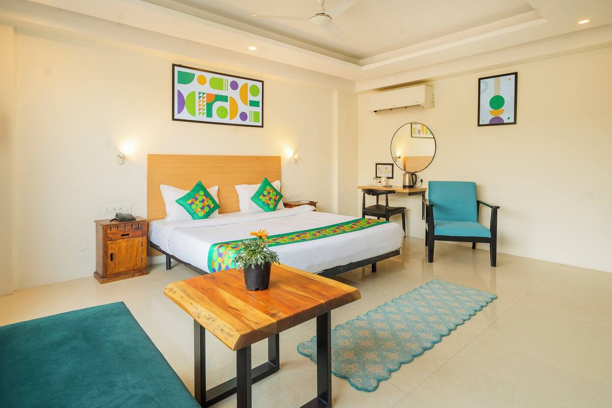 A hotel room with a bed, a desk, a chair, a small table, a potted plant, and a mirror.