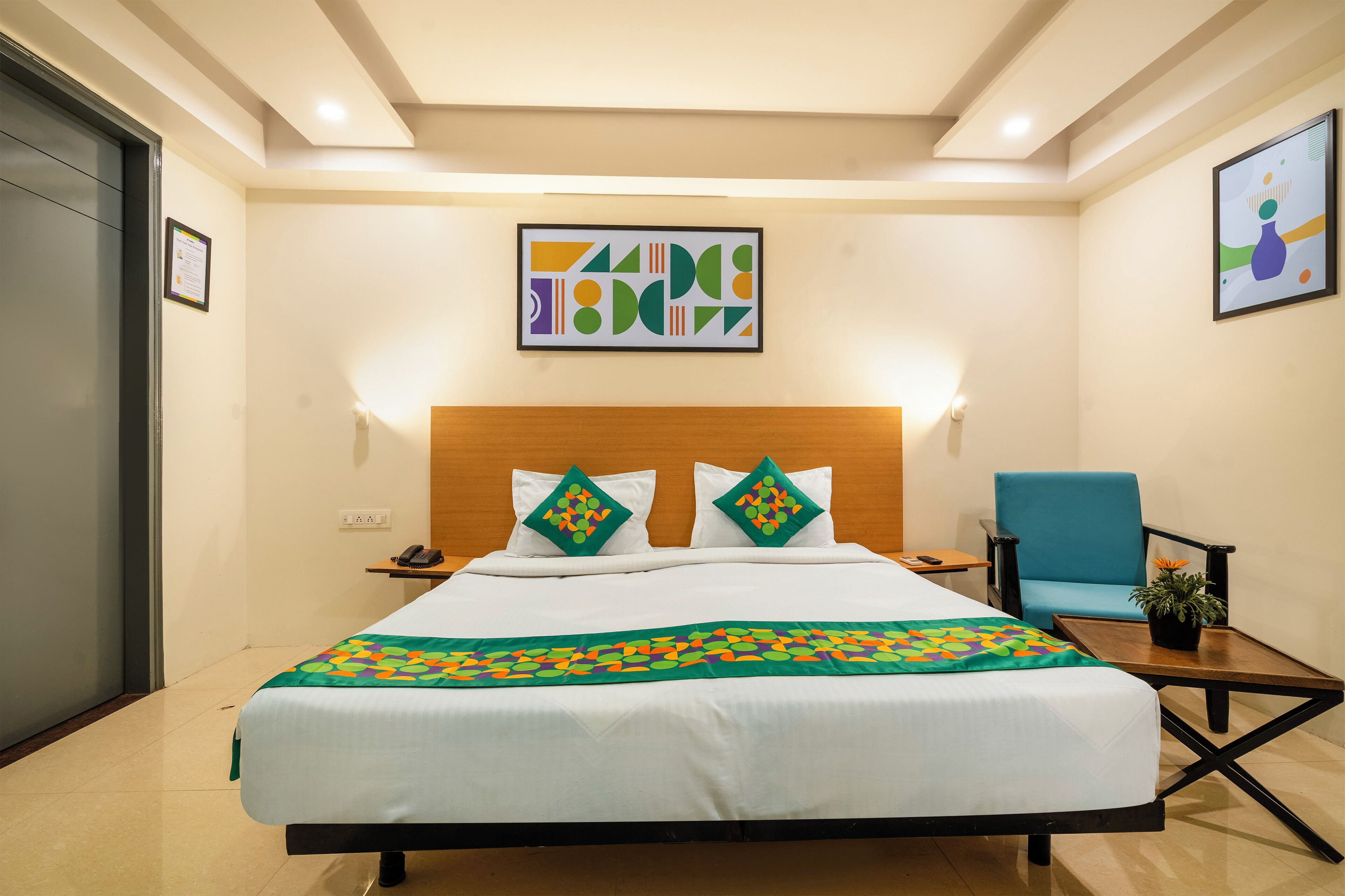 Una habitación de hotel con una cama, un sillón azul, una mesita con una planta y obras de arte en las paredes.