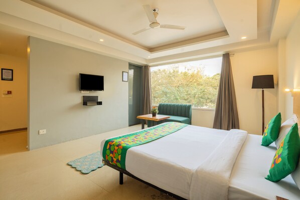 A hotel room with a bed, a television, a window with curtains, a ceiling fan, and a small table with a plant. - Treebo Twin Tree Suites (Gurugram)