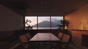 A dining area with a wooden table and chairs, a view of a mountain and lake through large windows, and a plant on the windowsill. - Good Fellows Resort (Kagoshima)