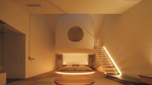 A modern hotel room with a minimalist design, featuring a central wooden table, a staircase with a wooden railing, and a circular skylight. - Good Fellows Resort (Kagoshima)