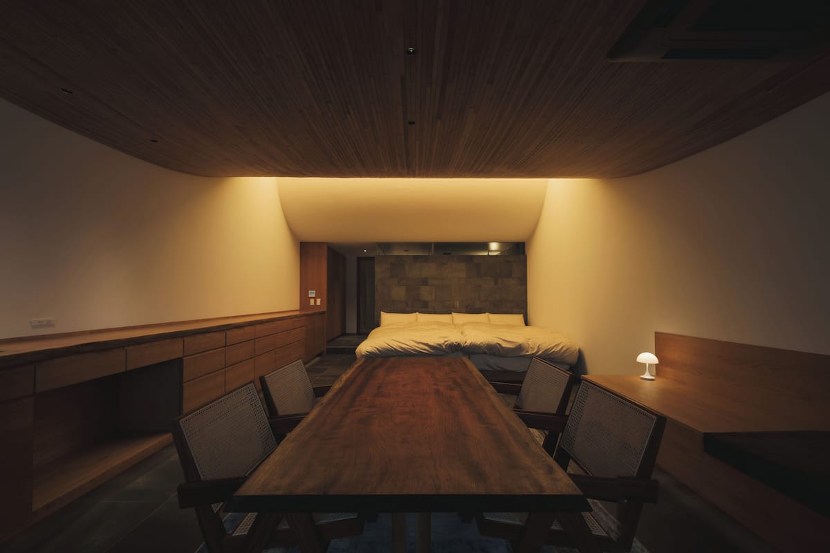 Un dormitorio moderno con techo de madera, una cama grande, una mesita de noche con lámpara y un escritorio de madera con sillas.