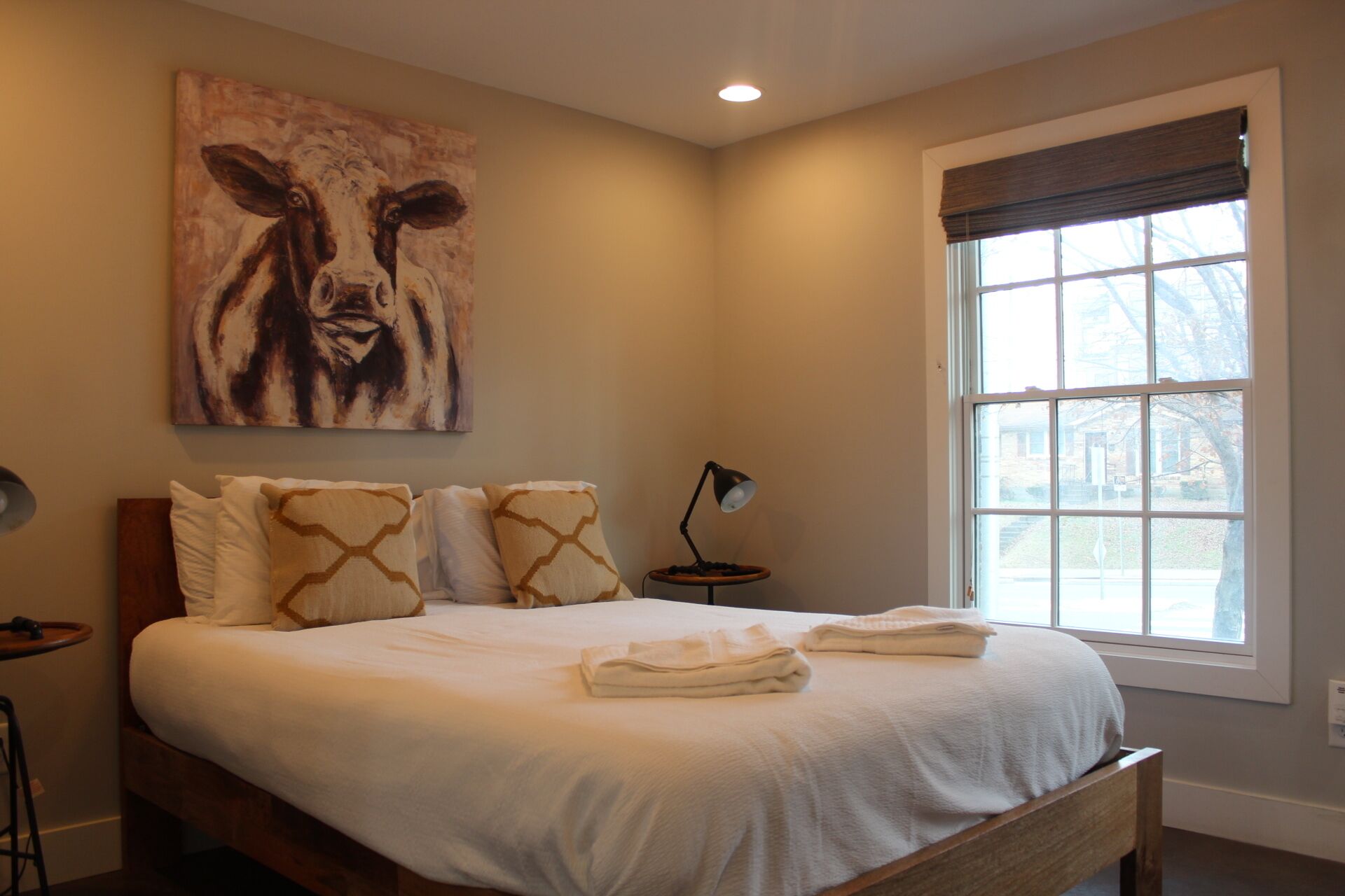 A neatly made bed with white linens and a brown headboard, a large abstract painting on the wall, and a window with blinds.