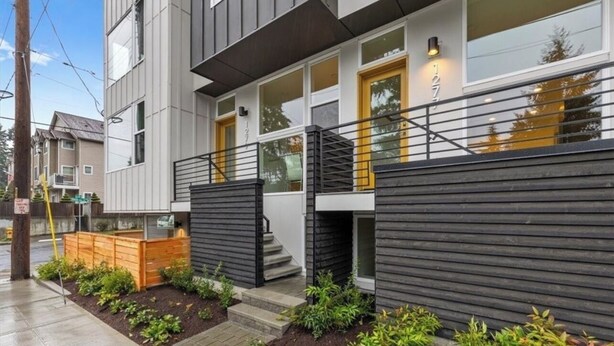 A modern two-story building with a flat roof, large windows, and a wooden fence.