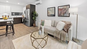A modern kitchen with a built-in oven and refrigerator, a dining area with stools, a living room with a sofa and a coffee table, and a large window providing natural light.