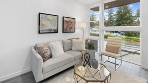 A modern living room with a sofa, a glass coffee table, and a chair. There are two framed artworks on the wall and a view of a balcony with a chair.