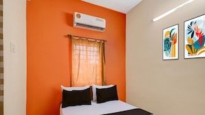 Une chambre avec un mur orange, un lit avec une literie noire et blanche, un climatiseur, une fenêtre avec un rideau à motifs et deux œuvres d’art encadrées accrochées au mur.