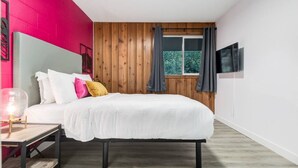 Une chambre à coucher avec un lit, une table de chevet, une fenêtre avec des rideaux et un téléviseur fixé au mur.