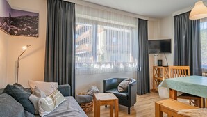 Un salon moderne comprenant un canapé, une petite table, un fauteuil et une table à manger avec des chaises. Un téléviseur est fixé au mur et il y a une fenêtre avec des rideaux.