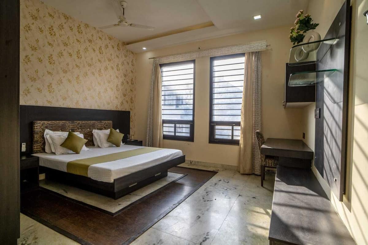 Un dormitorio con cama, mesitas de noche, escritorio y una silla. Hay dos ventanas con cortinas, un ventilador de techo y una estantería con una planta.