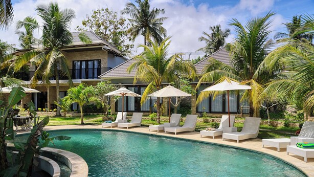 A pool area with lounge chairs and umbrellas, surrounded by palm trees and a building with a curved roof. - Kelingking Mesari Villa & Spa (Penida Island)