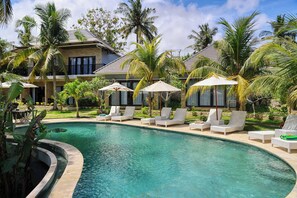 A pool area with lounge chairs and umbrellas, surrounded by palm trees and a building with a curved roof. - Kelingking Mesari Villa & Spa (Penida Island)