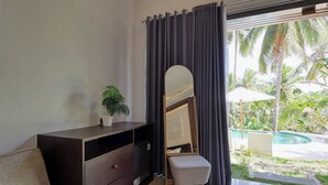 A bedroom with a dark wooden dresser, a small potted plant, a mirror, and a view of a swimming pool and tropical plants outside. - Kelingking Mesari Villa & Spa (Penida Island)