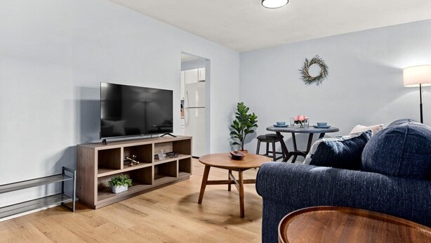 Un salon moderne doté d’un téléviseur à écran plat, d’un meuble de rangement en bois, d’une table basse ronde en bois, d’un canapé bleu et d’une petite table à manger avec des chaises.