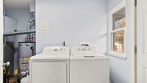 Une buanderie équipée de deux lave-linge et deux sèche-linge à chargement frontal, tous deux de couleur blanche, d’un chauffe-eau et d’une fenêtre donnant sur les arbres.