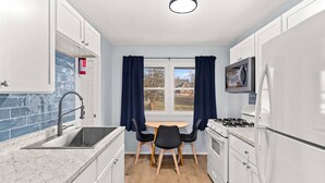 Une cuisine compacte avec des meubles blancs, un évier, une cuisinière et un four à micro-ondes. Il y a une fenêtre avec des rideaux, une petite table avec deux chaises et un extincteur accroché au mur.