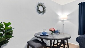 Un coin repas avec une table ronde, de la vaisselle bleue, un vase avec des fleurs, une couronne décorative accrochée au mur et un lampadaire.