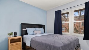 Une chambre à coucher avec un lit, une table de chevet, une petite plante et une fenêtre avec des rideaux.