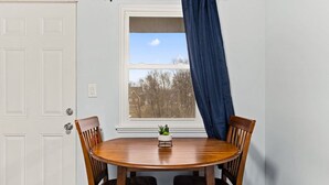 Ein Essbereich mit einem runden Holztisch, vier Stühlen, einem kleinen Topfpflanzen und einem Fenster mit Blick auf Bäume und Gebäude.