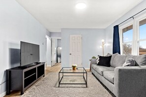 A modern living room with a flat-screen TV, a grey sofa, and a glass coffee table. - Home Suite Home-M: Minutes to Downtown & P Heights (Peoria)