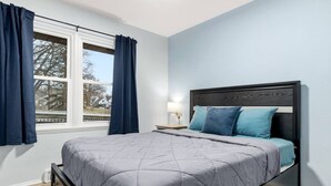 Une chambre à coucher avec un lit, une table de chevet, une fenêtre avec des rideaux et une vue sur l’extérieur.