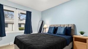 Quarto com cama, mesa de cabeceira, luminária e janela com cortinas.