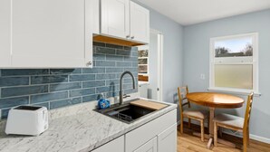 Uma cozinha com armários brancos, painel de azulejos azuis, pia de aço inoxidável, torradeira e mesa de jantar de madeira com cadeiras.