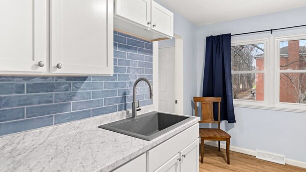A kitchen with white cabinets, a blue-tiled backsplash, a sink, and a chair near a window.
