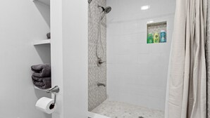 Un espace douche équipé d’un pommeau de douche, d’un porte-serviettes avec des serviettes, d’un support à papier toilette et d’une étagère contenant des articles de toilette.