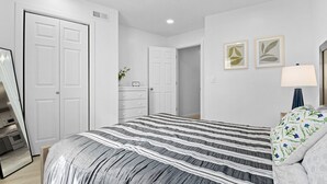Une chambre à coucher avec un grand lit, deux portes blanches, une commode, un miroir et deux tableaux encadrés accrochés au mur.