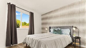 Une chambre à coucher avec un lit, des rideaux, une table de chevet, une lampe et une vue sur l’extérieur.