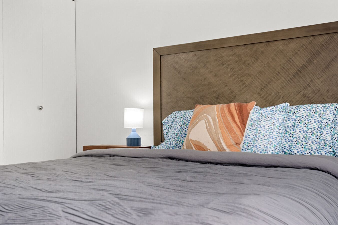 A neatly made bed with a grey comforter and patterned pillows, a bedside table with a lamp, and a wooden headboard.