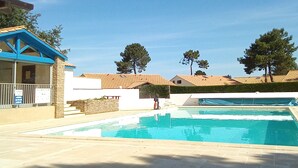Une piscine avec une terrasse carrelée, un bâtiment doté d’un toit bleu et des arbres en arrière-plan.