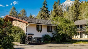 Un edificio de dos pisos con fachada de piedra y techo inclinado, rodeado de árboles y montañas al fondo.
