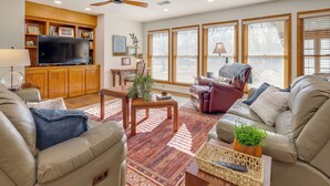 A living room with a flat-screen TV, wooden cabinets, a ceiling fan, and large windows with blinds. - Fish, Hike & Relax: Heber Springs Family Home (Heber Springs)
