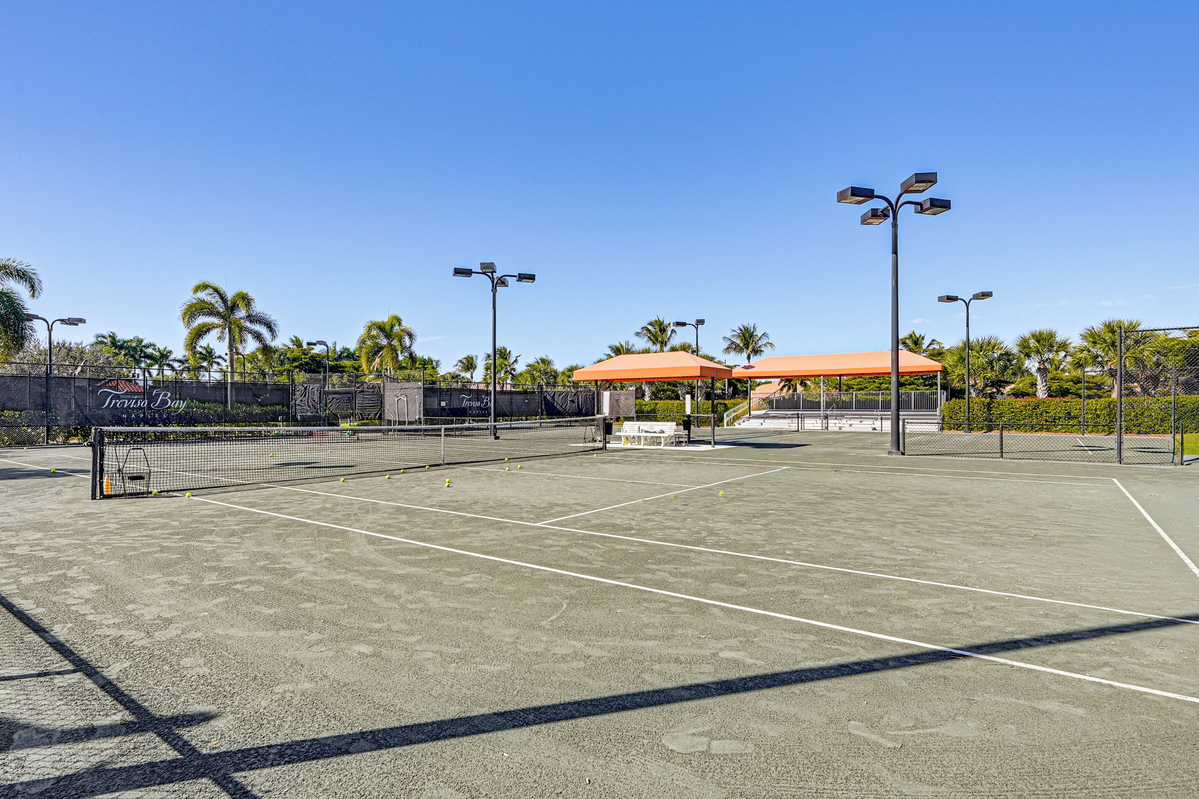 An outdoor tennis court with a net, surrounded by palm trees and a covered seating area.