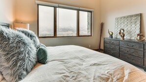 A bedroom with a large window, a bed with a fluffy pillow, a dresser with decorative items, and a wall-mounted artwork. - Luxe Eden Gem w/ Hot Tub: 6 Mi to Powder Mountain! (Eden)