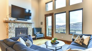 Ein Wohnzimmer mit Kamin aus Stein, einem flachbildfernseher darüber, einem großen Sofa, einem Couchtisch und großen Fenstern mit Blick auf eine verschneite Landschaft.
