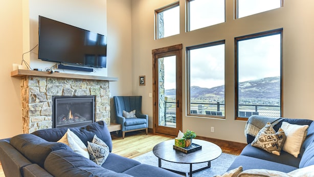 A living room with a stone fireplace, a flat-screen TV mounted above it, a large sofa, a coffee table, and large windows offering a view of a snowy landscape. - Luxe Eden Gem w/ Hot Tub: 6 Mi to Powder Mountain! (Eden)