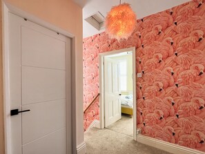 A hallway with a white door, a mirror reflecting a room with pink flamingo wallpaper, and a hanging light fixture. - A great pet friendly cottage just a five minute drive from Staithes. (Hinderwell)