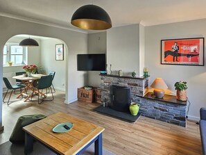 A living room with a fireplace, a television mounted on the wall, and a dining area with chairs and a table. - 3 bedroom accommodation in Newquay (Newquay)