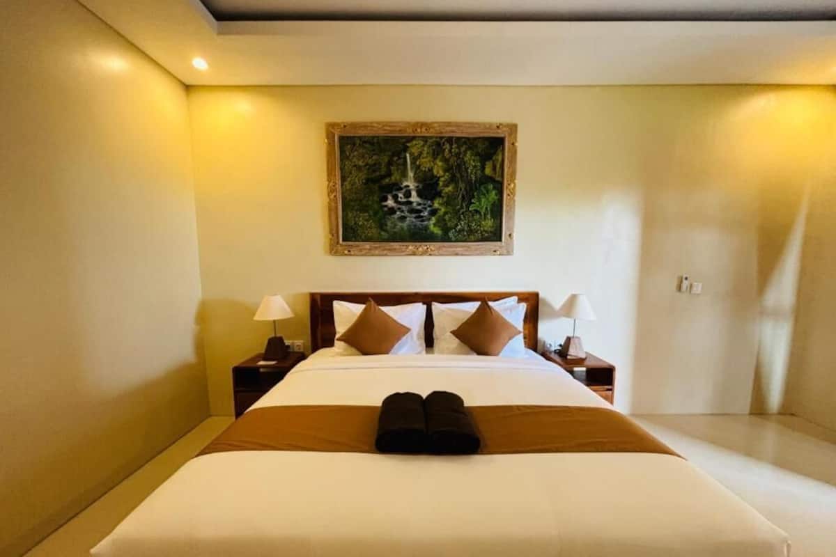 Ein Hotelzimmer mit einem großen Bett, zwei Nachttischen, einem Bild an der Wand und einem Paar zusammengerollter Handtücher auf dem Bett.