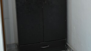 Une armoire de couleur sombre, à deux portes et dotée d’un tiroir, située dans un coin avec un sol carrelé.