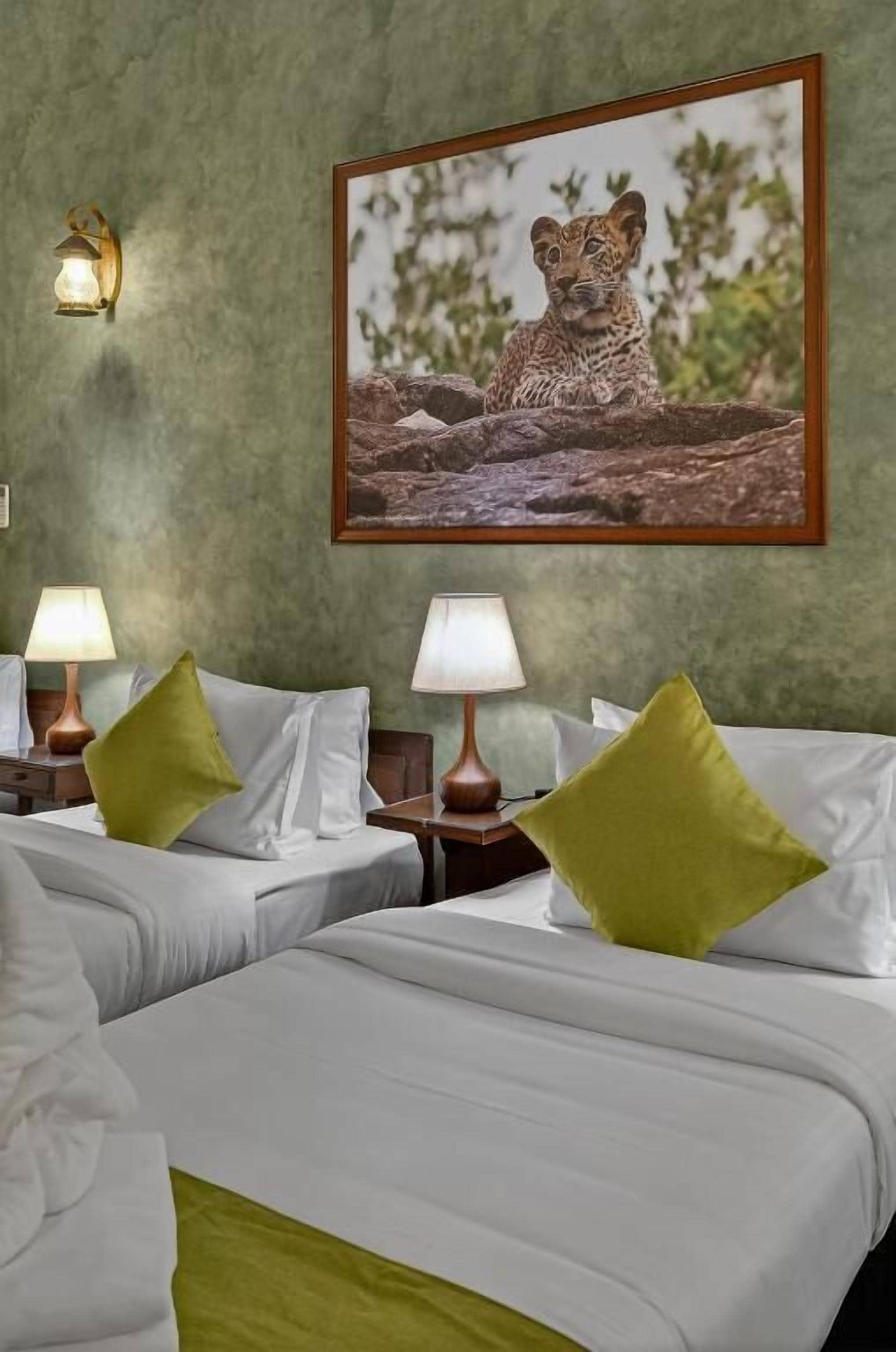Habitación de hotel con dos camas, almohadas verdes, un cuadro enmarcado de un leopardo y lámparas fijadas a la pared.
