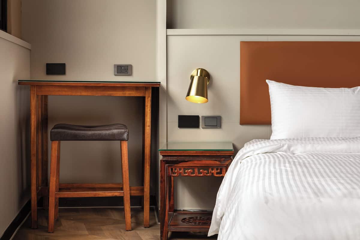 A hotel room with a bed, a wooden nightstand with a lamp, a small table, and a bench.