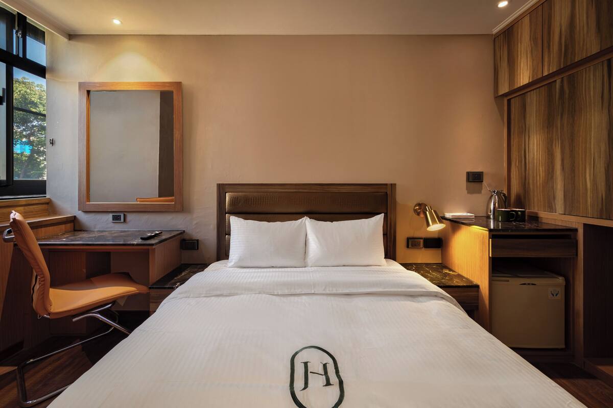 A hotel room with a bed, a desk, a chair, a small refrigerator, and a mirror on the wall.
