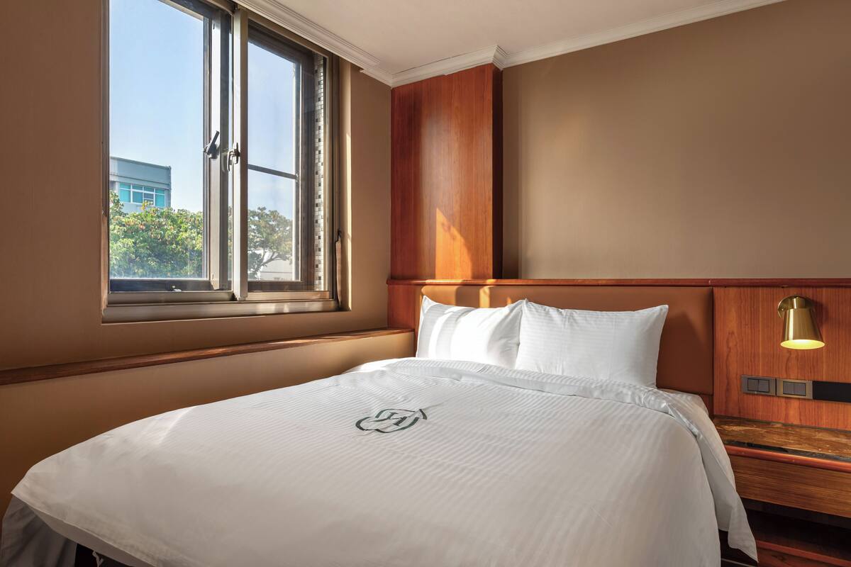 A hotel room with a large bed, a window with a view of buildings, a wooden headboard, and a bedside lamp.