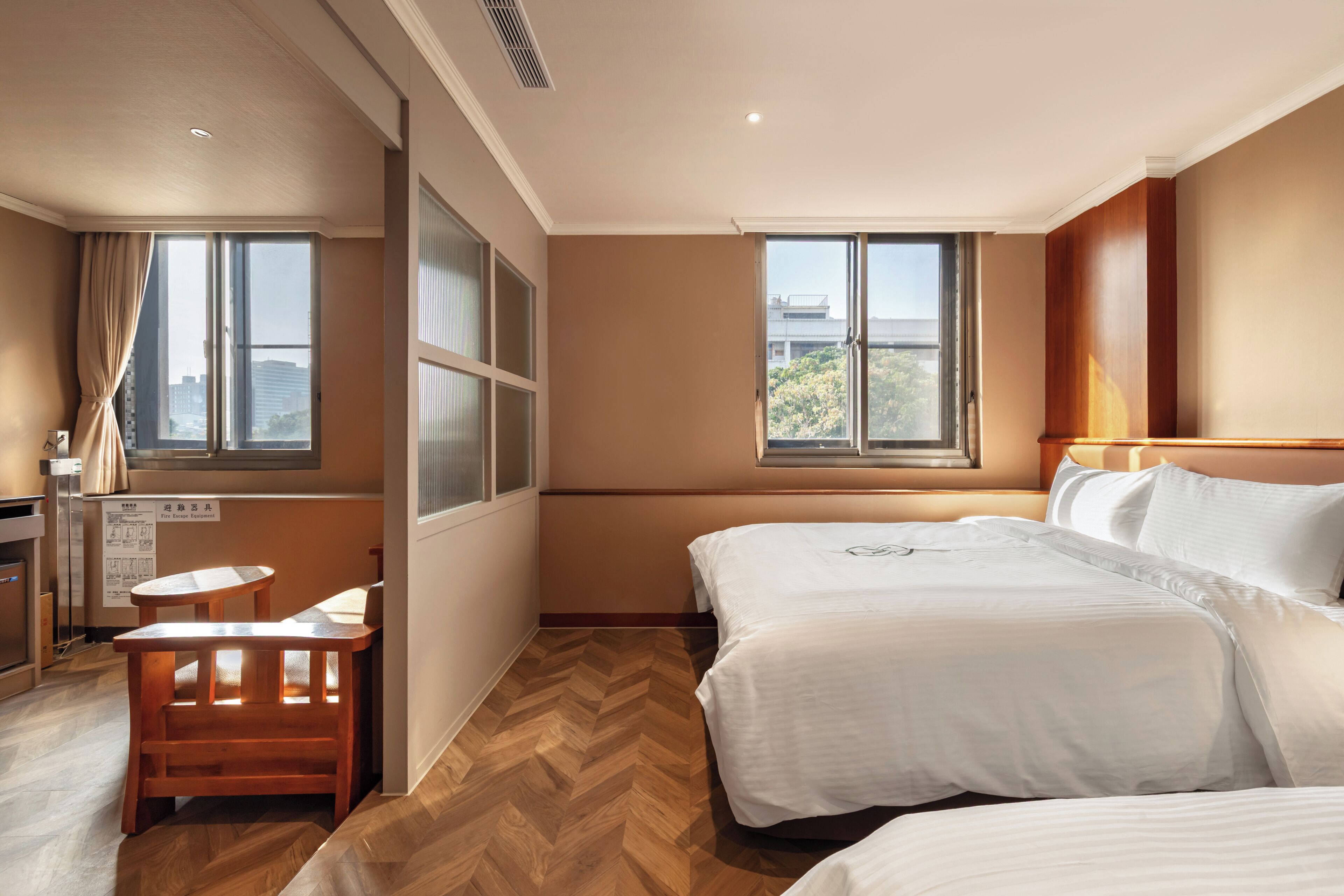 Ein Hotelzimmer mit zwei Betten, einem Schreibtisch, einem Stuhl und großen Fenstern mit Blick auf die Stadt.